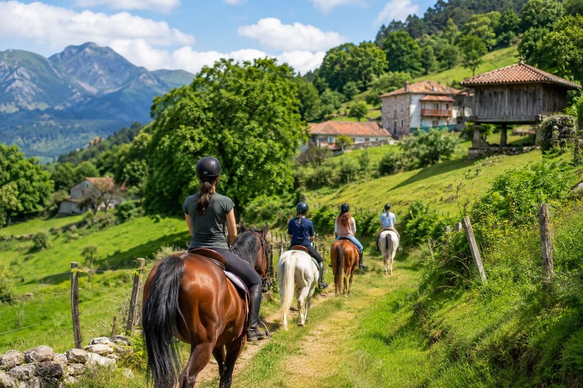 Grupo de jinetes recorriendo un camino rural entre prados verdes y hórreos en un paisaje de montaña de Asturias