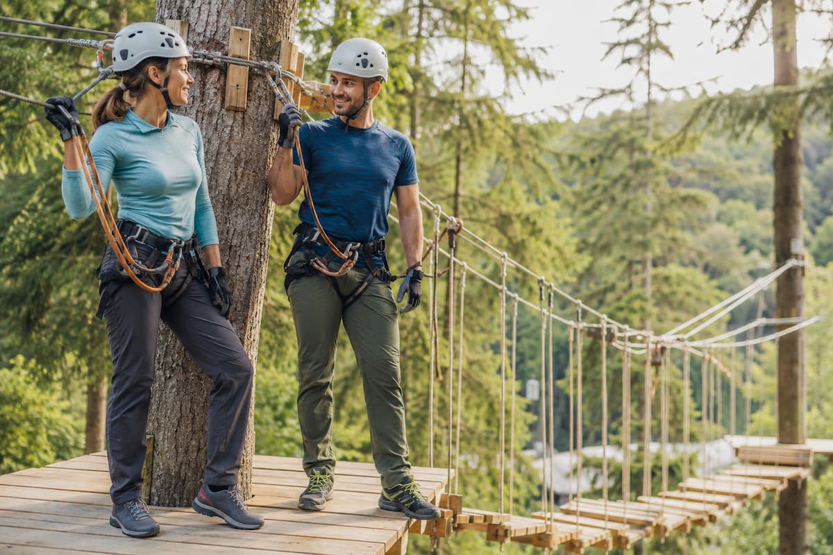 Pareja equipada con casco, arnés, ropa técnica y zapatillas de montaña junto a un circuito de puentes colgantes.