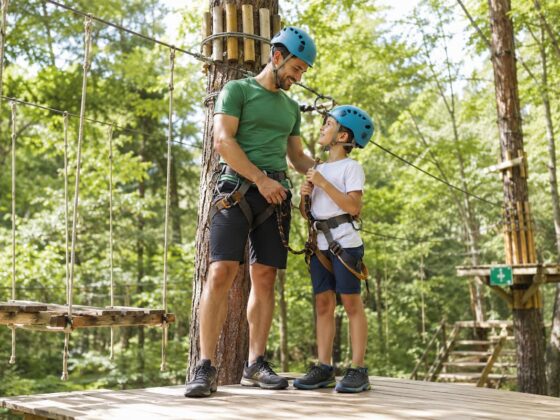 Padre e hijo con casco y arnés en una plataforma de un parque multiaventura, vestidos con ropa deportiva y calzado cerrado.