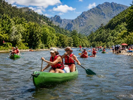 Dos personas con chalecos rojos remando en una canoa verde durante el Descenso del Sella, con más participantes y montañas al fondo.